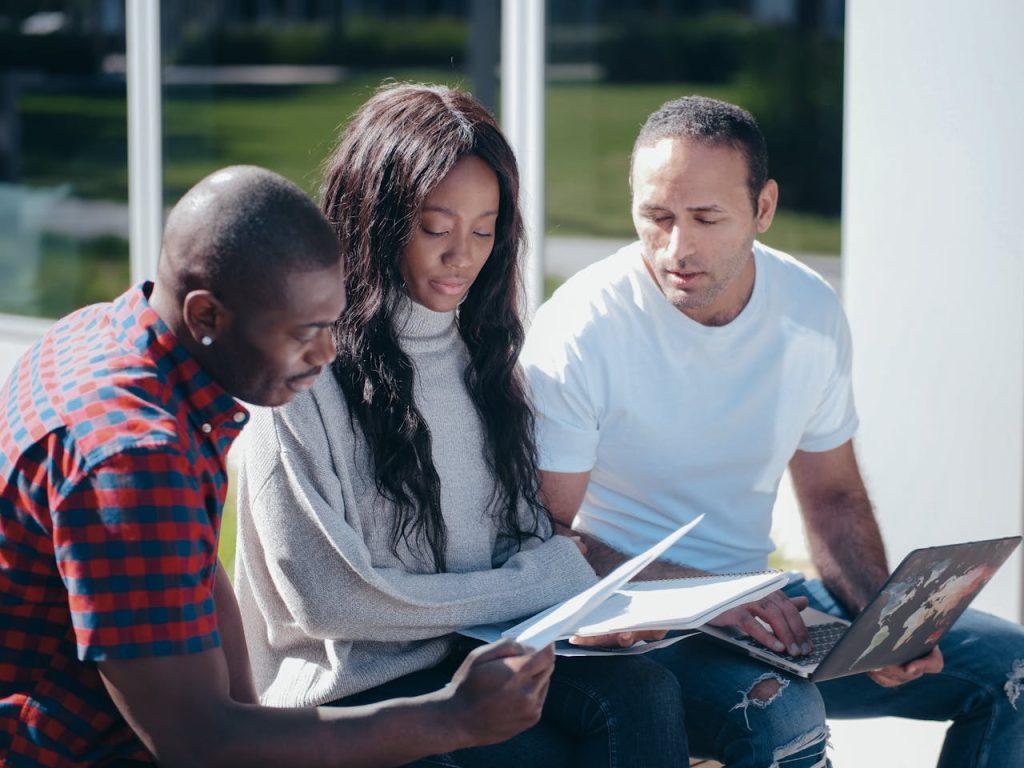 Three colleagues collaborating outdoors, discussing documents and using a laptop.